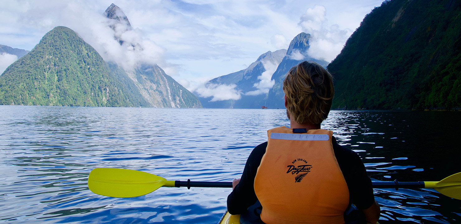 Student kayaking in a bay with tall, cloud-obscured islands in the background.